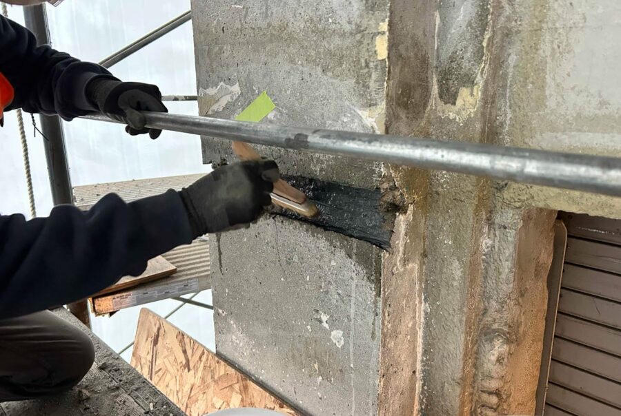 A person wearing gloves applies black sealant or adhesive to a crack in a concrete wall, using a brush. Scaffolding and construction materials are visible in the background.