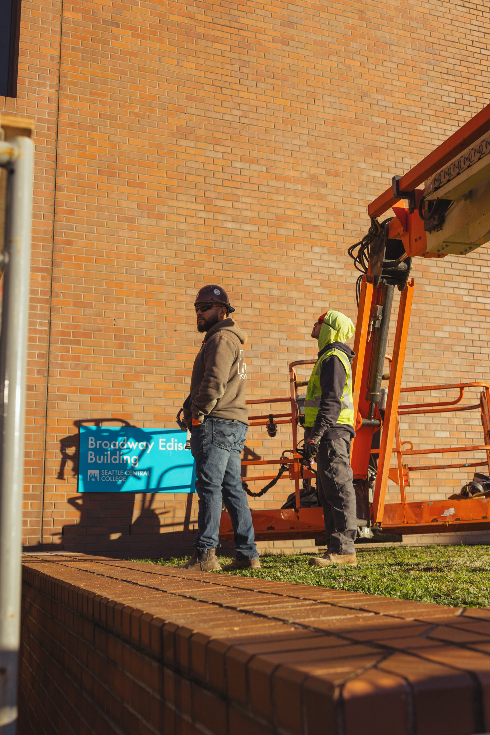 Two construction workers stand near a cherry picker lift in front of a brick building with a blue sign reading Broadway Edison Building in sunlight.