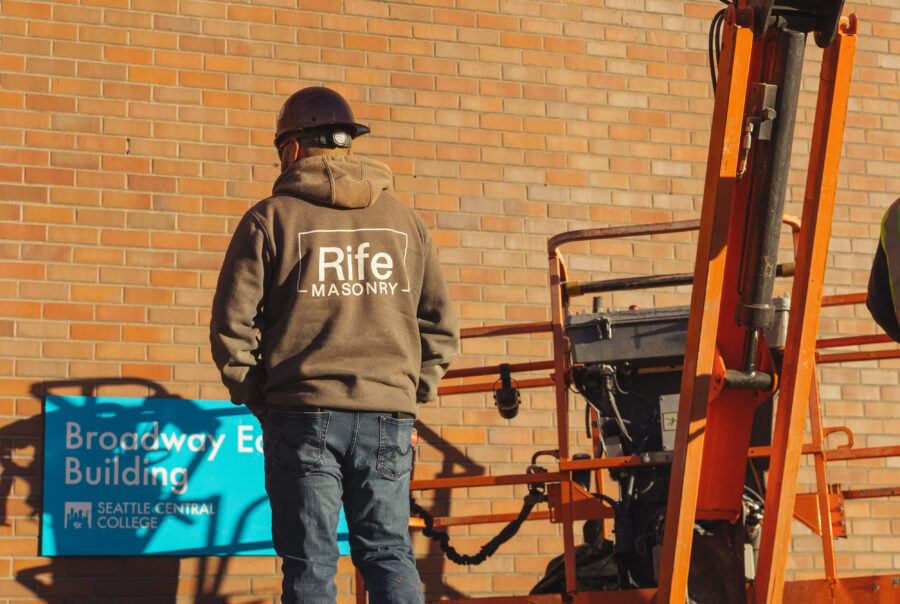 A construction worker wearing a hard hat and a Rife Masonry jacket walks toward a brick wall near an orange lift. A sign reading Broadway Edison Building, Seattle Central College is visible on the wall.