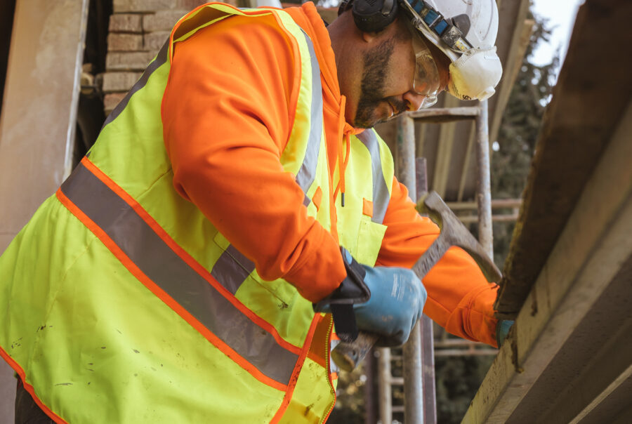 A construction worker in a neon safety vest, orange hoodie, gloves, helmet, and ear protection works with scaffolding at a building site. Trees and construction materials are visible in the background.