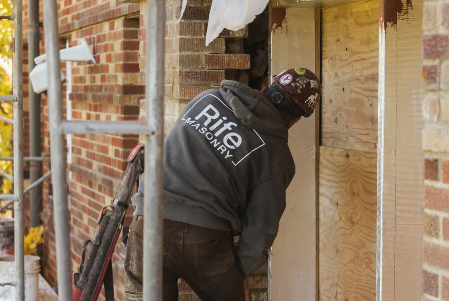 A construction worker wearing a Rife Masonry sweatshirt works on a brick buildings exterior near a boarded-up doorway, surrounded by scaffolding and construction tools.