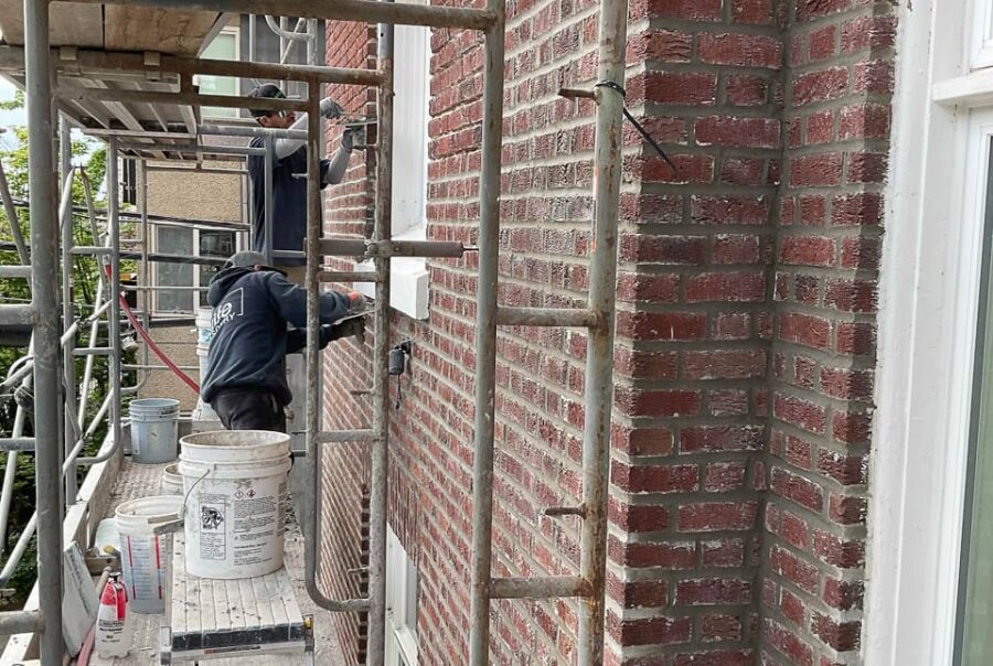 Two workers on scaffolding repair the brick facade of a building. Buckets and tools are on the platform, and the area is surrounded by metal scaffolding and a white-framed window is visible on the right.