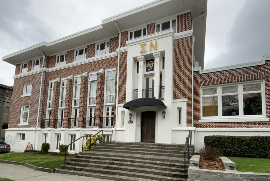 A large brick building with white trim and tall windows, featuring Greek letters ΣΝ above the main entrance, wide front steps, and a small lawn in front.