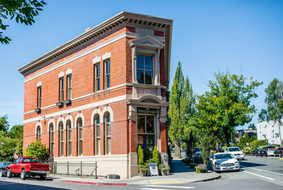 A historic red brick building with arched windows and a Windermere sign sits on a sunny street corner lined with cars, trees, and a sidewalk.