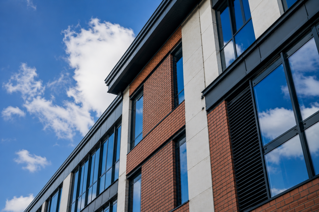 Modern building with large windows and a mix of red brick and white panels, reflecting a blue sky with scattered clouds. Viewed from a low angle.
